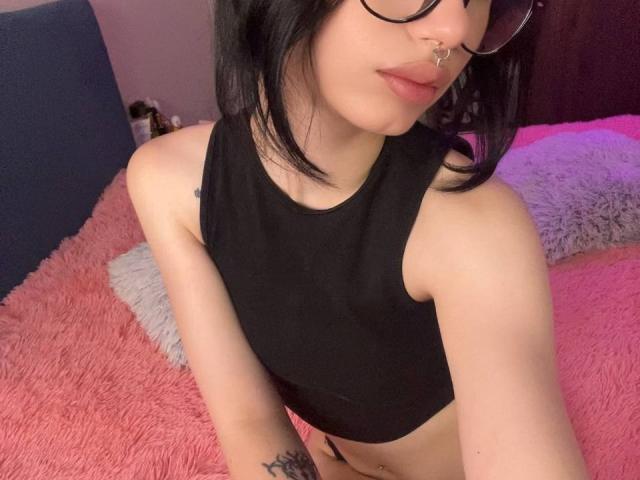 GlitchDoll with black hair, glasses, nose ring, black sleeveless top, exposing her bare tits and a small tattoo on her right arm, sitting on a pink bed.