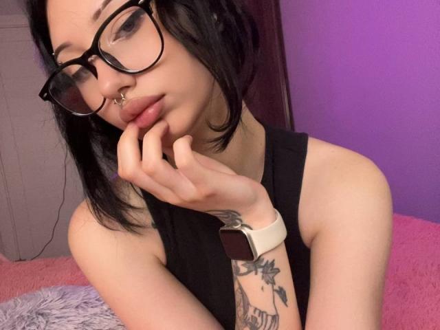 GlitchDoll with black hair, glasses, nose ring, and a watch, touching her chin, showing a tattoo on her arm, in a purple and pink setting.