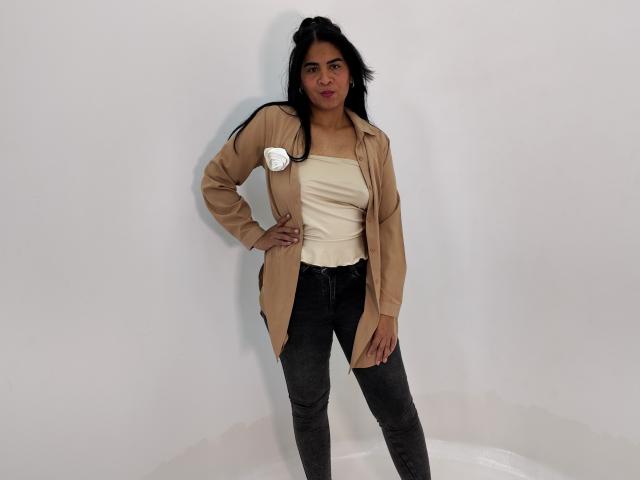 Rhouss stands with a hand on her hip, wearing a beige jacket, white top, and black jeans. She has a white flower on her jacket and a confident expression.