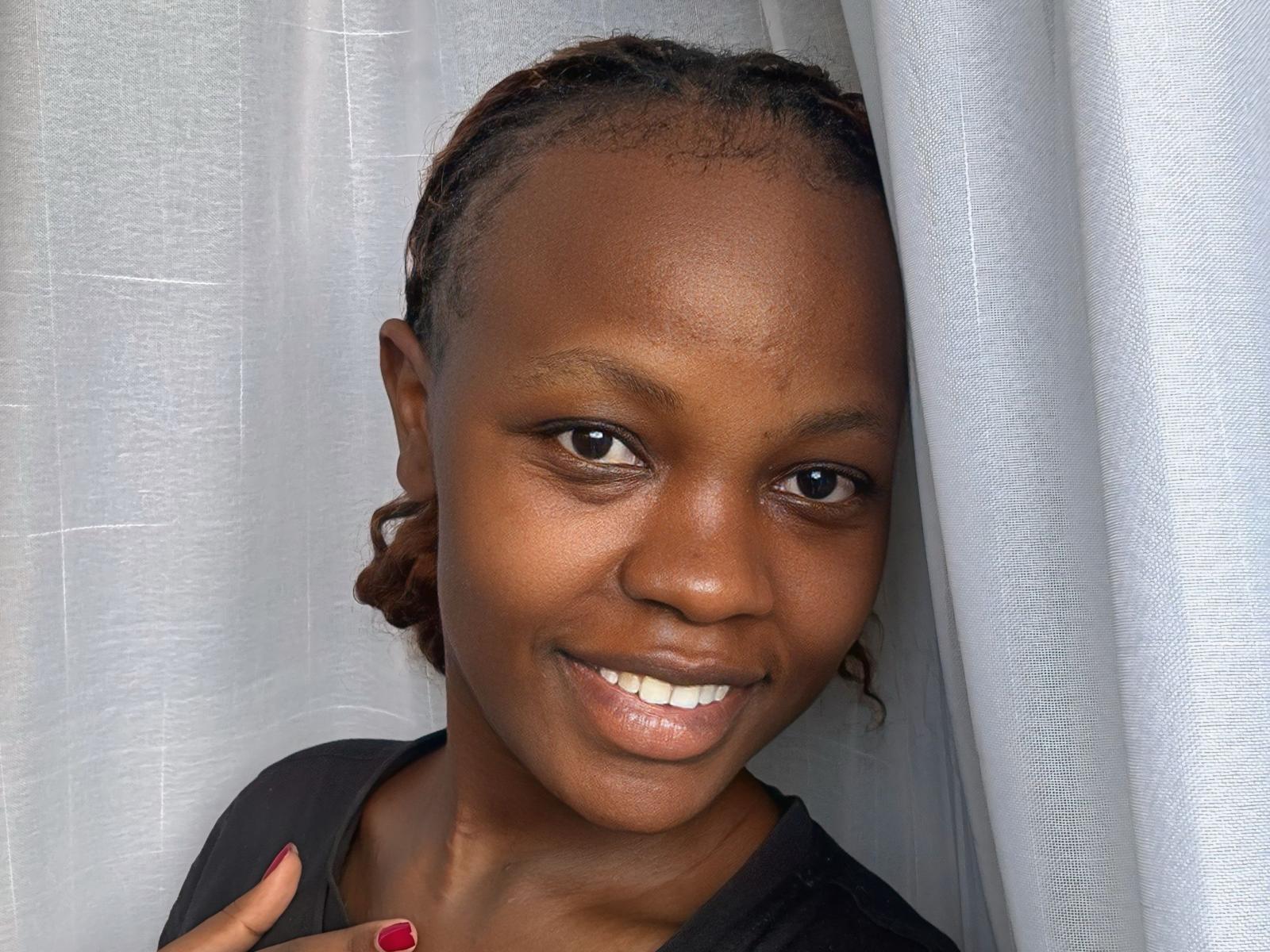 A photo of SweetheartCandy with dark brown skin, braided hair, and a smile showing white teeth. She's wearing a black shirt, with red nails, standing in front of white curtains.