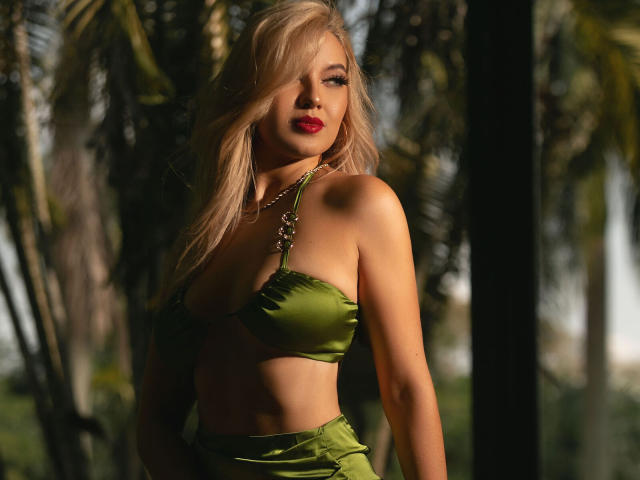 AlanaMonroe, com cabelo loiro, batom vermelho, sutiã de cetim verde e calças de cintura alta combinando, em pé sob a luz do sol, mostrando seus seios e um colar, com um leve sorriso..