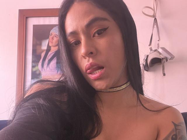 StellaBeckker with long black hair, a choker, and winged eyeliner, showing her shiny pink lips. Her tits are partially visible. A portrait of another woman is in the background.