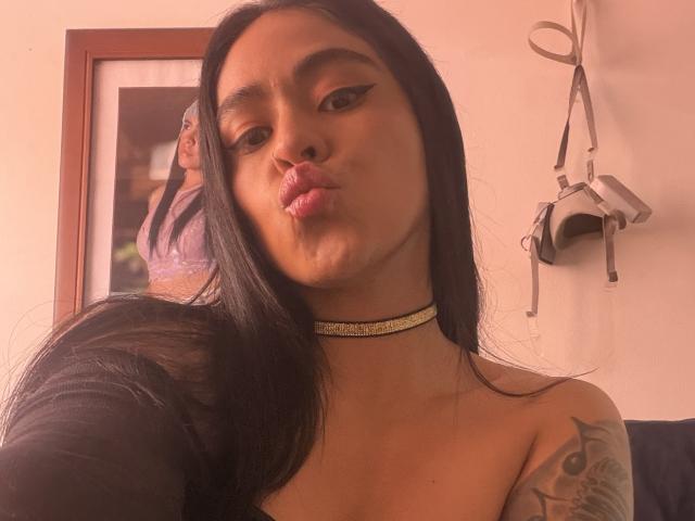 StellaBeckker with long black hair, wearing a choker, pouting her lips, showing a tattoo on her right shoulder, in a selfie. Boobs are visible but not the focus.