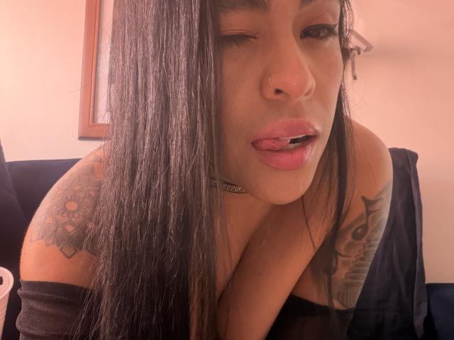 StellaBeckker has long black hair, a nose ring, and is winking with her tongue out. She has tattoos on her shoulder and arm, and is wearing a choker.