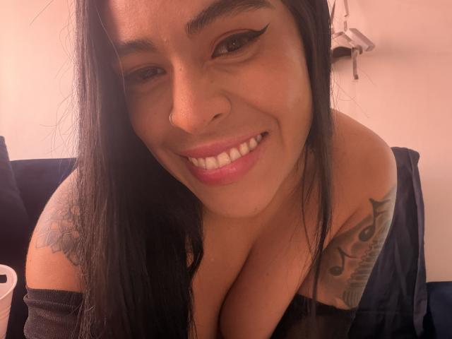StellaBeckker heeft lang zwart haar, een neusring, zwarte eyeliner, roze lippenstift en een tatoeage op haar rechter schouder. Ze glimlacht en haar borsten zijn zichtbaar, ze draagt een zwarte top..