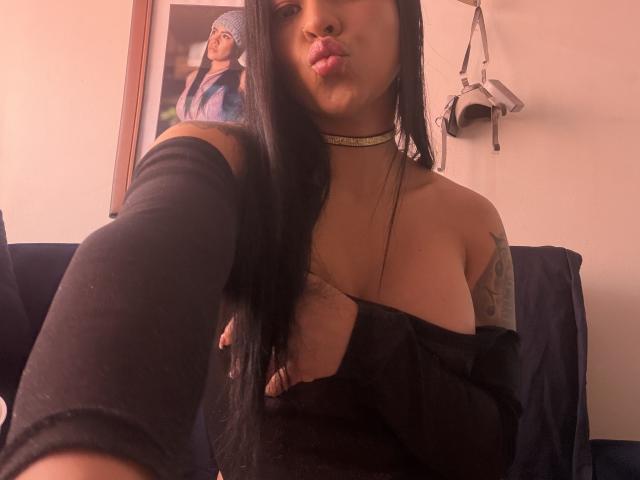 StellaBeckker with long black hair, pouty lips, black off-shoulder top, gold choker, covering small tits, tattooed arm, black sleeve, white wall, hanging bag, framed photo in background.