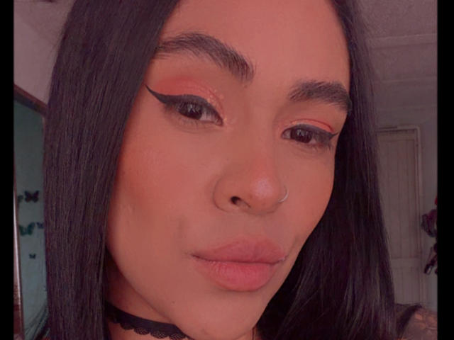 StellaBeckker has dark straight hair, a nose ring, and winged eyeliner. Her pinkish-orange makeup and black choker are visible. Her plump pink lips are slightly parted.