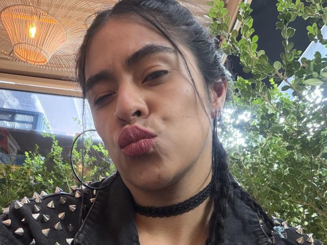 StellaBeckker, met volle, roze lippen, een zwarte choker en een leren jas met studs, maakt een grappig gezicht. Ze heeft donker haar, donkere wenkbrauwen en een kleine blauwe plek op haar wang..