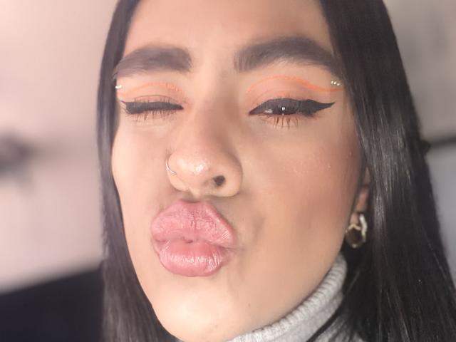 StellaBeckker with one eye closed, wearing orange eyeliner, gold piercings, nose ring, and pink glossy lips, black straight hair, and a gray turtleneck, blowing a kiss.