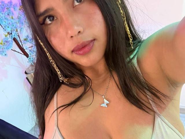 GemmaBellini with long brown hair, wearing gold chain earrings and a silver butterfly necklace, showing her tits in a white tank top, and a slight smile.