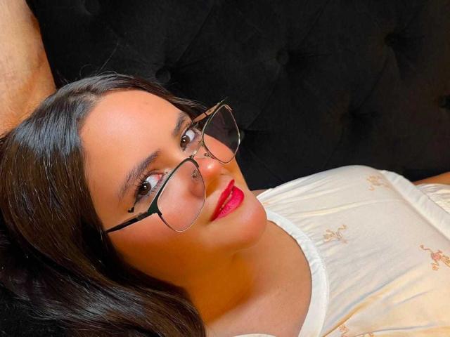 EmilyColsson heeft een medium bruine huid, zwart haar, rode lippenstift, een bril en een neusring, en ligt op een zwarte, gestoffeerde bank, gekleed in een witte top met bloemenprint..