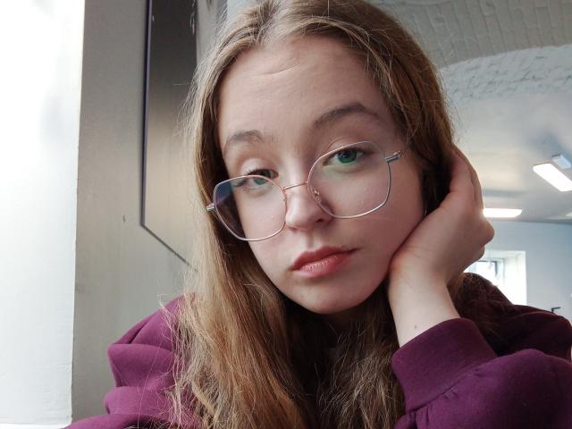 MandiBouie, con gafas, cabello castaño, labios rosados y una sudadera morada, con la mano apoyada en la mejilla, con expresión seria, ojos verdes, labios ligeramente fruncidos, en interiores, luz natural, informal..