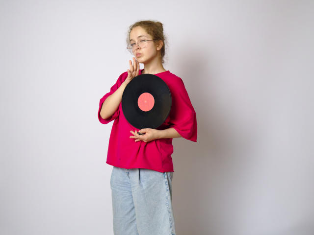 MandiBouie, con i capelli ricci, gli occhiali e una camicia rosa, tiene un disco in vinile nero sopra il seno, indossa una gonna grigia ed è in piedi contro un muro bianco..