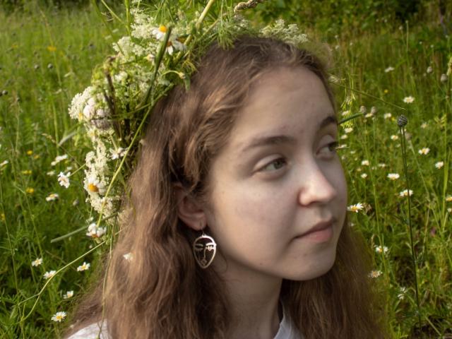 MandiBouie, avec ses cheveux bruns ondulés, une couronne de fleurs blanches, des yeux verts, un léger sourire, portant une boucle d'oreille en forme de cœur argentée, se tient dans un champ herbeux avec des marguerites..