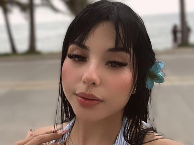 GraceClarke has wet black hair with a blue flower, a nose piercing, and is wearing a striped halter top, showing off her tits with a subtle, confident smile.