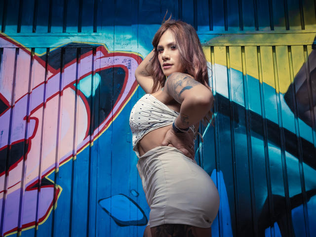 AzhunaStar met roze haar, tatoeages, een wit crop top en een strakke witte rok, staat zijwaarts, met haar hand op haar heup, waarbij ze haar achterkant laat zien en naar de camera kijkt. Een kleurrijke muur met graffiti..