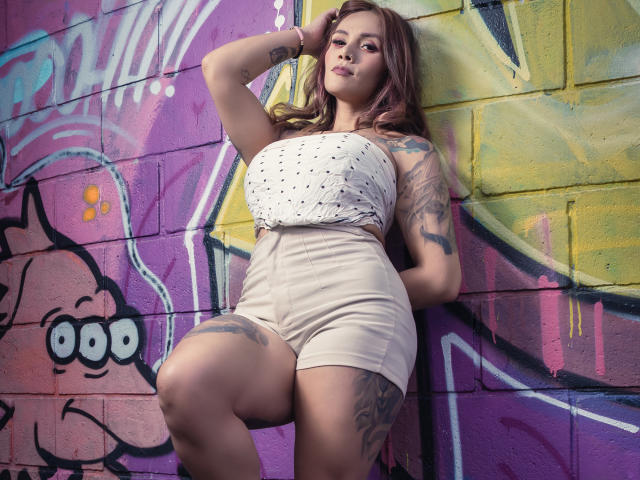 AzhunaStar with big tits, wearing a white dotted top and beige shorts, leaning against a colorful graffiti wall, showing tattoos on her arm and thigh.