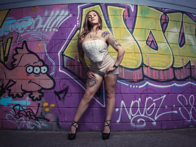 AzhunaStar with tattoos, wearing a white strapless top, beige shorts, black high heels, standing against colorful graffiti, hands on hips, legs apart, looking up, confident.