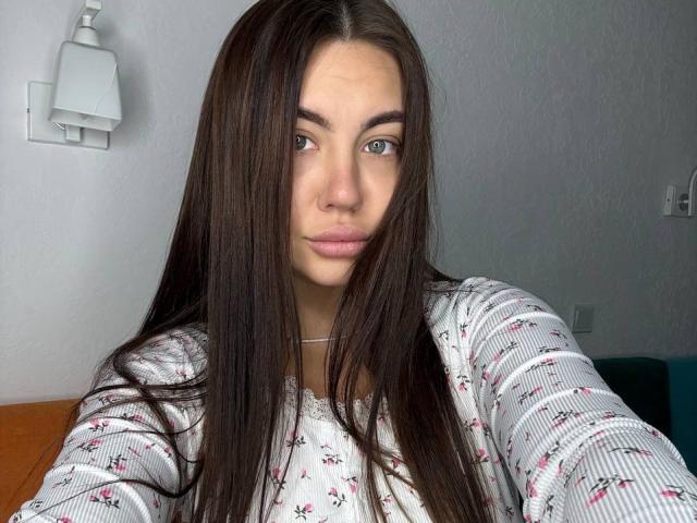 Ines Gueningsman hat langes, glattes, braunes Haar, helle Haut und blaue Augen. Sie trägt ein weißes, geblümtes Hemd und blickt direkt in die Kamera, wobei sie einen neutralen Gesichtsausdruck hat..