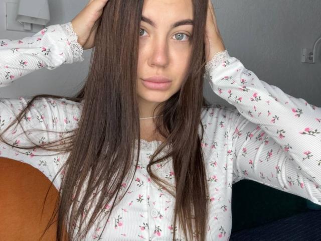 InesGueningsman with long brown hair, wearing a white floral shirt, hands in hair, blue eyes, pink lips, sitting, holding an orange pillow.