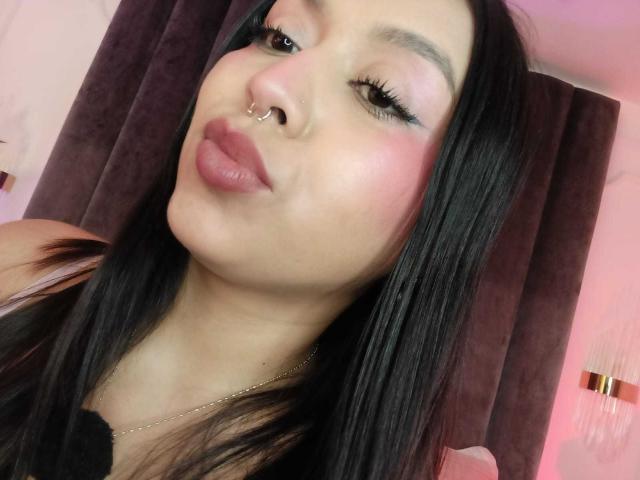ZammySanz tiene el pelo largo y negro, un piercing en la nariz, colorete rosa y labios rosados y gruesos, lleva una blusa negra con un collar dorado y mira a la cámara..