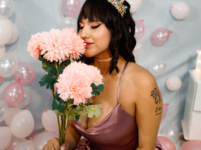 HanaJoud with pink flowers, wearing a shiny pink dress, choker, and tiara, smelling roses, tattoo on right shoulder, pink and white balloons in background.