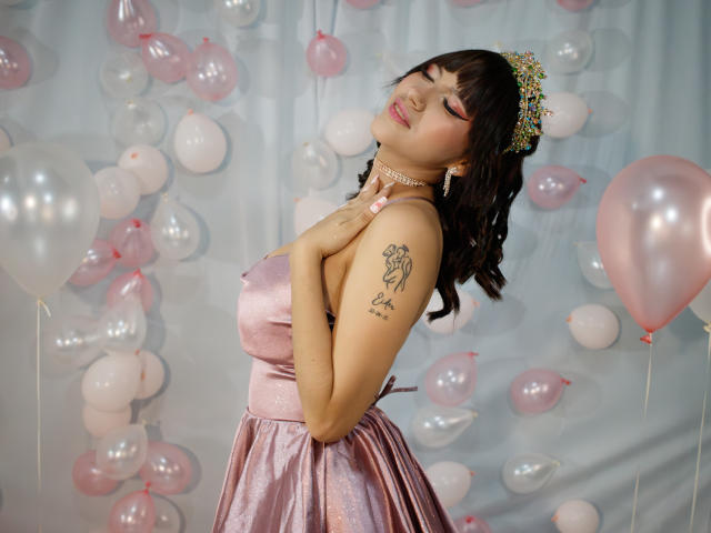 HanaJoud with pink satin dress, floral headpiece, pink and white balloons, tattoo on right arm, hands on chest, eyes closed, pink lipstick, pink nails, pink choker.