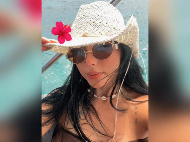 GraceClarke with black hair, wearing a straw hat with a red flower, round sunglasses, white earbuds, and a shell necklace, showing her tits in a sunlit boat.