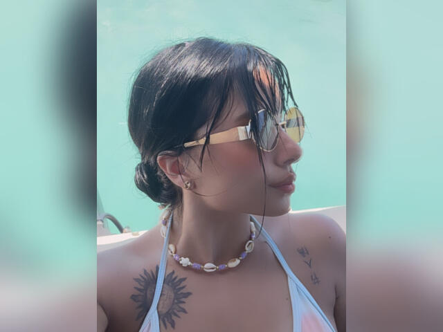 GraceClarke with wet black hair, wearing yellow sunglasses and a white bikini, has sun tattoos on her chest and a seashell necklace. Her tits are visible.