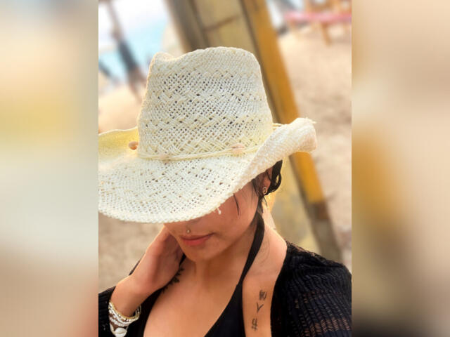 GraceClarke with tan skin, wearing a white crochet hat, black halter top, and black knit cardigan, has a nose ring, tattoo on right side, and a bracelet on her left wrist.