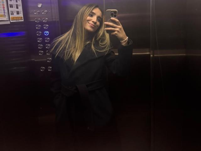 AdharaCooperr, avec de longs cheveux blonds, souriante, portant un manteau noir, prend un selfie dans un ascenseur sombre, tenant un smartphone avec une coque rose..