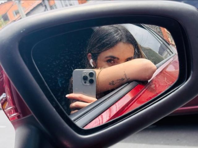 AdharaCooperr's reflection in a car side mirror, showing her brown eyes, black hair, earbuds, tattoo on her right arm, and holding a gray iPhone. Red car visible.