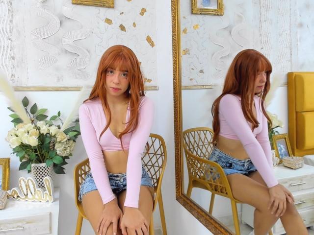 AmelieBenson with red hair, pink long-sleeve top, and denim shorts, sitting on a golden chair, reflection in mirror, showing her small tits and cute ass.