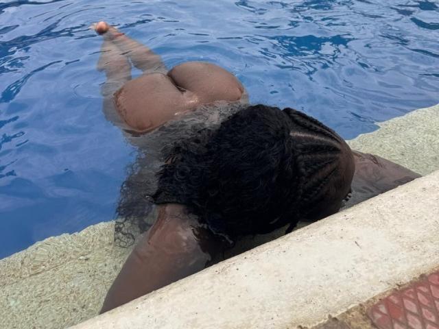 SamyLorenss's ass is in the blue water, face down with braided black hair, arms resting on a concrete edge, legs slightly apart, toes pointed.