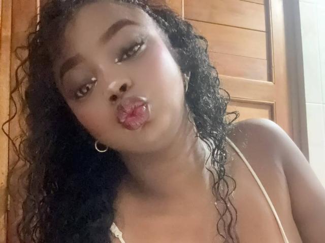 SamyLorenss with dark curly hair, hoop earrings, and pink lipstick pouting her full, glossy, wet-looking lips. She's wearing a white spaghetti strap top, showing her small, round tits.