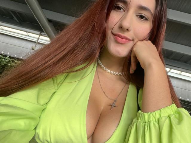 JuicyLana mit langen braunen Haaren, in einem grünen Kleid, das tiefe Ausschnitte zeigt, mit einer Hand auf der Wange, lächelnd, trägt eine silberne Kreuzkette und einen Choker, unter einem Metalldach..