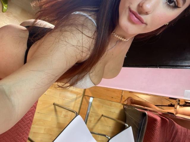 JuicyLana, con lunghi capelli castani, indossa uno slip nero e un reggiseno bianco, mostrando il seno e i glutei in un selfie, sorridendo, con uno sfondo di una parete rosa e un pavimento di legno..