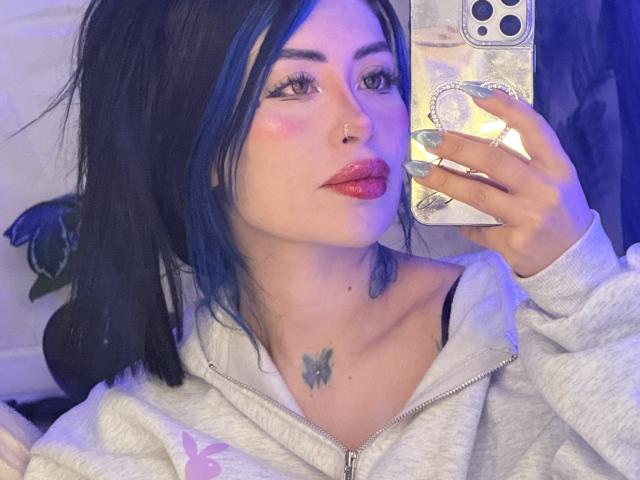 Kkole with black hair, blue highlights, nose ring, and butterfly tattoo on chest, wearing grey hoodie, holding phone with long silver nails, pink lipstick, and blue-purple lighting.