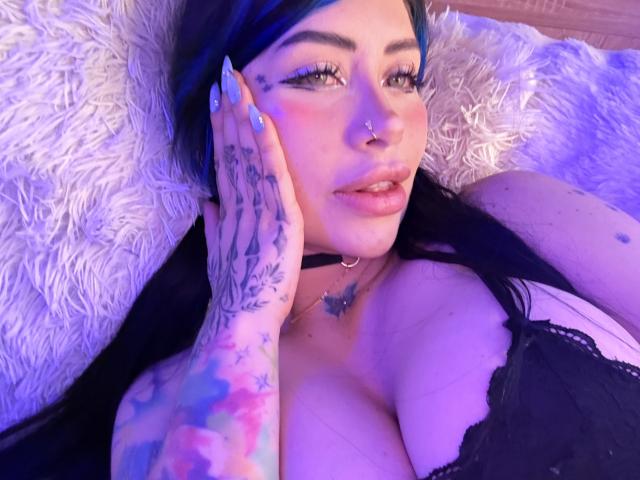 Kkole, met blauwe highlights, eyeliner en een neusring, ligt op een pluizig wit oppervlak en laat haar kleurrijke tatoeages zien, haar grote borsten in zwart kant, en een vlinderketting..