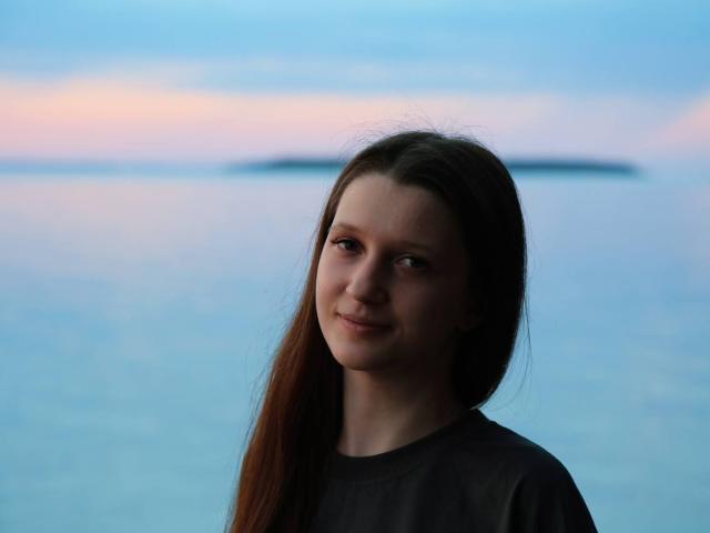 Joetta Savka, con lunghi capelli castani, piccoli seni e un leggero sorriso, indossa una camicia scura e si trova accanto a una calma distesa d'acqua blu al tramonto..
