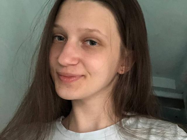 Joetta Savka, com pele clara, olhos castanhos e cabelos longos castanhos, sorrindo levemente, vestindo uma camisa branca, com um nariz discreto e sem seios, mamas ou vulva visíveis..
