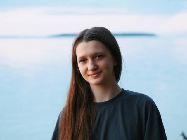 Joetta Savka, mit langen, braunen Haaren, lächelnd, trägt ein dunkles Hemd, hat blaue Augen und helle Haut. Sie steht an einem ruhigen, blauen Gewässer und blickt direkt in die Kamera..