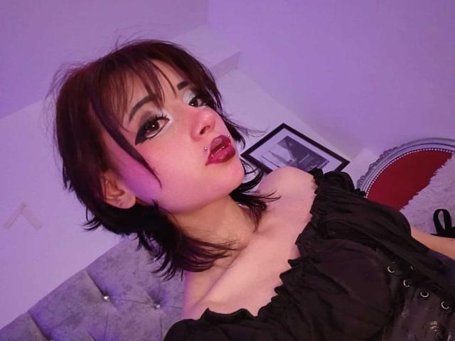 ViolettClay has dark brown hair, heavy black eyeliner, pinkish-purple eye shadow, and red lipstick. She's wearing a black off-shoulder top, showing her small tits. Purple room.
