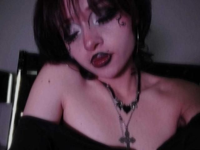 ViolettClay with dark makeup, red lipstick, black off-shoulder top, black necklace, cross pendant, and black hair, looking down with a slight frown, showing bare shoulders and chest.