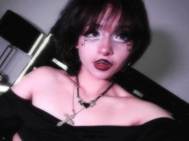 ViolettClay with black hair, white face paint, and dark lipstick, tongue out, black off-shoulder top, necklace, and earrings, in a slightly blurry photo.
