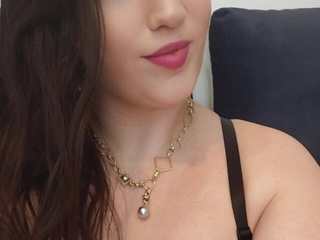 VanessaSquirtt with pink lipstick, dark wavy hair, gold chain necklace, black strap top, showing her neck and upper chest, smiling slightly. Her tits are partially visible.