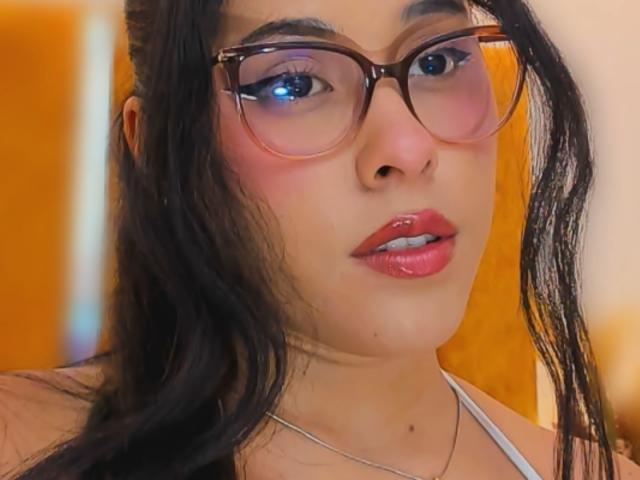 MarilynHills has glossy pink lips, shiny black hair, and wears thick brown glasses with blue eyeliner. Her nose is small, and she has a silver necklace.