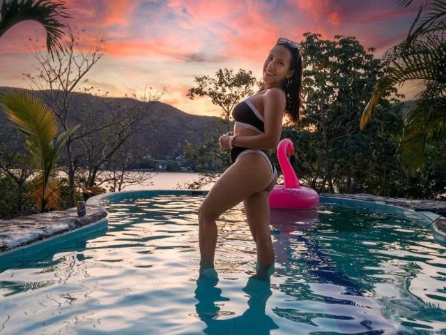 Jade Foster, avec sa peau bronzée et son corps voluptueux et sexy, porte un maillot de bain noir et blanc et se tient dans une piscine avec un flotteur en forme de flamant rose, avec un coucher de soleil en arrière-plan..