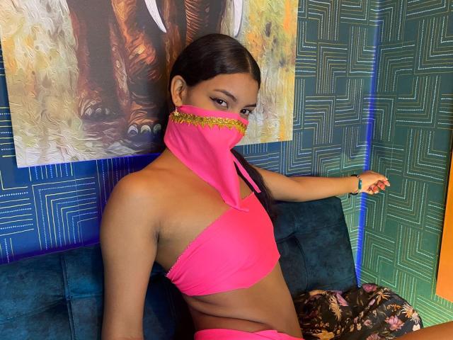 LalilaBrunette, dark-skinned with a flat stomach, wearing a pink halter top and matching mask with gold trim, sitting on a blue couch, showing her tits.