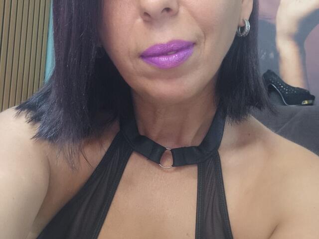 LauraSin with purple lipstick, black halter top, small ring at neck, black shoulder-length hair, hoop earrings, smiling, showing her tan skin and bare shoulders, looking sexy.
